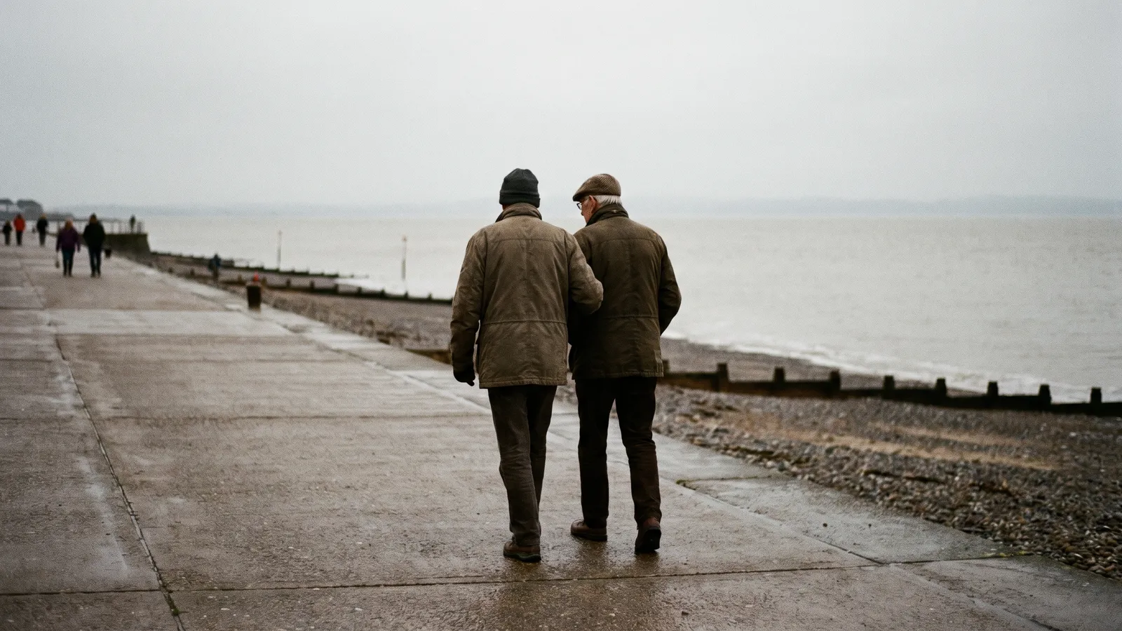 Two older men in winter coats walking slowly side by side along a grey seafront promenade, seen from behind