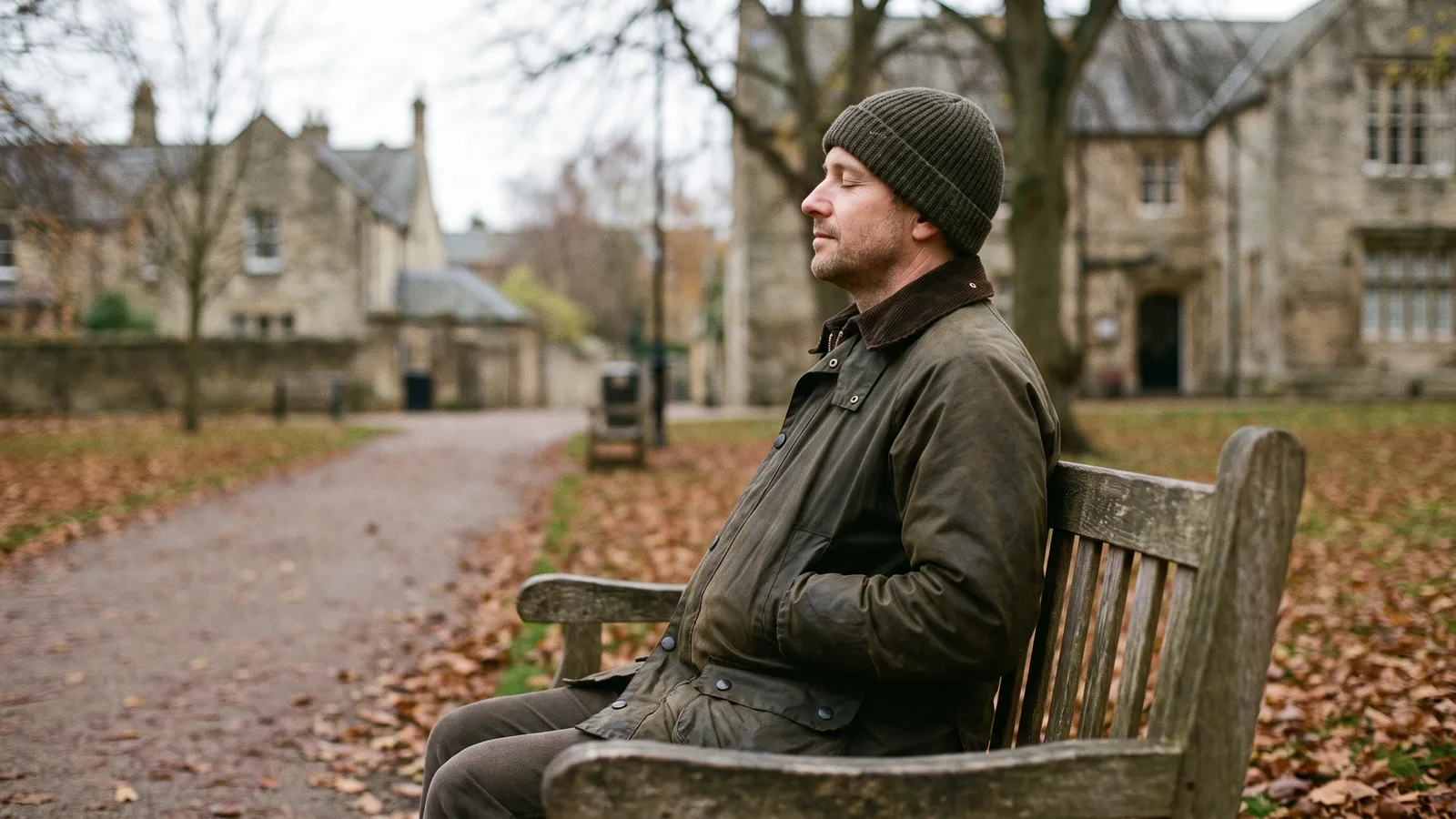 A man in a waxed jacket sitting on a wooden park bench in autumn with his eyes closed, breathing slowly